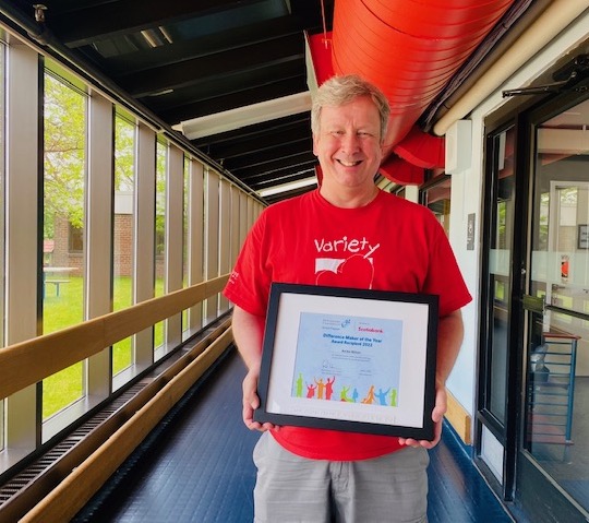 Homme d’âge moyen aux cheveux blonds courts portant un t-shirt rouge. Il tient un certificat encadré du prix Personne qui fait une différence de l’année.